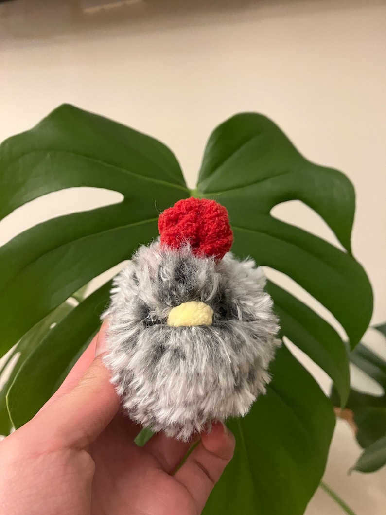 Puede incluir: Un peque&ntilde;o pollo de crochet gris y blanco con una cresta roja de crochet. El pollo tiene un pico amarillo y ojos negros.