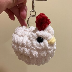 Puede incluir: Llavero de pollo blanco tejido a crochet con un pomp&oacute;n rojo en la cabeza y ojos negros. El llavero tiene un anillo plateado y un clip plateado.