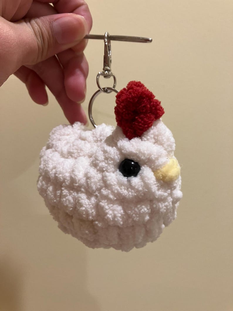 Puede incluir: Llavero de pollo blanco tejido a crochet con un pomp&oacute;n rojo en la cabeza y ojos negros. El llavero tiene un anillo plateado y un clip plateado.
