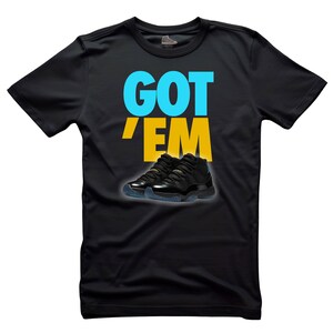 May include: Black t-shirt with the text "GOT 'EM" in blue and yellow. Below the text is an image of black and gold sneakers with blue soles. The shirt is made of a soft material.