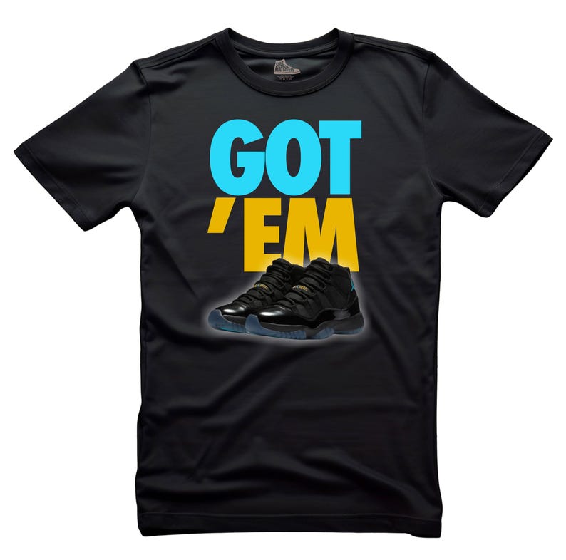 May include: Black t-shirt with the text "GOT 'EM" in blue and yellow. Below the text is an image of black and gold sneakers with blue soles. The shirt is made of a soft material.