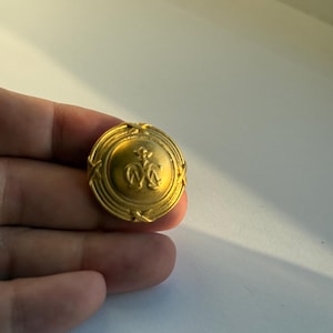 Pode incluir: Um botão dourado com um emblema em relevo no centro. O botão é redondo com uma borda decorativa. O emblema parece ser um design estilizado. O botão é segurado em uma mão.