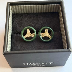May include: A pair of cufflinks in a black box. The cufflinks feature a green steering wheel design with gold-colored accents. The box is labeled "HACKETT MAYFAIR". The interior of the box is lined with a striped pattern.
