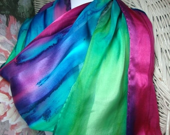 May include: A silk scarf with a gradient of colors, including purple, blue, green, and pink. The scarf is folded and draped over a white wicker chair.