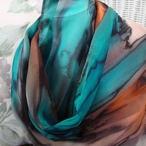 May include: A flowing silk scarf with a watercolor design. The scarf features shades of teal, brown, and orange, creating a dynamic and artistic pattern. The fabric is draped, showcasing its texture and the interplay of colors. A stylish accessory.