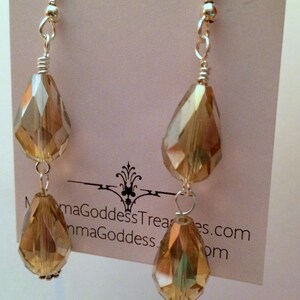 May include: A pair of dangle earrings with two teardrop-shaped crystals. The crystals are a light amber color and have a faceted design. The earrings are made with silver-toned metal.