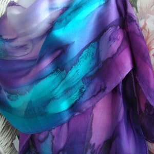 May include: A silk scarf with a vibrant watercolor design. The scarf features a blend of colors, including shades of purple, teal, and blue. The fabric is draped, showcasing the flow and texture of the material. The scarf is a fashion accessory.