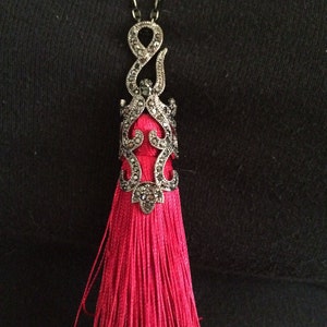 May include: A red tassel necklace with a silver filigree pendant. The pendant is adorned with black stones.