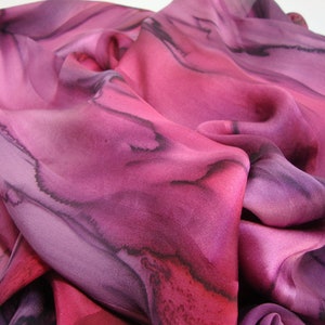 May include: A pink and purple silk scarf with black abstract designs. The scarf is made of a soft, flowing fabric and has a delicate, hand-painted look.
