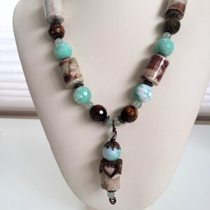 May include: A handmade necklace with brown, green, and turquoise beads. The necklace features cylindrical and round beads, and a heart-shaped pendant. The necklace is displayed on a white mannequin.