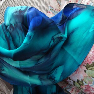 May include: A teal and blue silk scarf with abstract watercolour-like designs. The scarf is folded and draped over a floral patterned cushion.