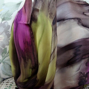 May include: A silk scarf featuring a flowing design in shades of purple, green, and brown. The scarf has a watercolour-like effect with abstract shapes and lines.