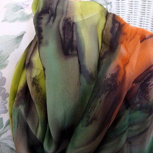 May include: A green, orange, and black silk scarf with abstract brushstrokes. The scarf is folded and draped over a white wicker chair.