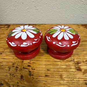 May include: Two red, hand-painted wooden cabinet knobs. Each knob features a white daisy design with a yellow centre and green leaves. The knobs are on a wooden surface.