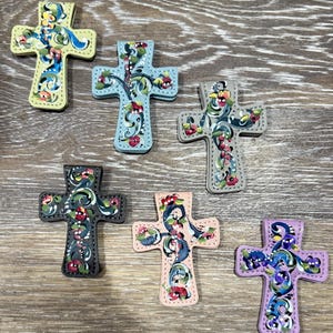 May include: A collection of seven decorative crosses with floral designs. Each cross features a different color scheme, including yellow, blue, gray, black, pink, and purple. The crosses are adorned with painted flowers and vines, creating a handcrafted appearance.