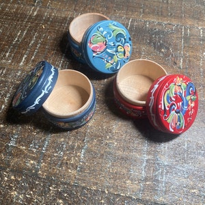 May include: Three small, round wooden boxes with lids. One is blue, one is red, and one is dark blue. Each lid is decorated with a floral design in various colors. The boxes are open, revealing the natural wood interior.