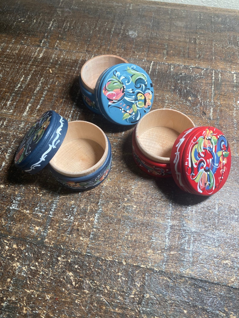 May include: Three small, round wooden boxes with lids. One is blue, one is red, and one is dark blue. Each lid is decorated with a floral design in various colors. The boxes are open, revealing the natural wood interior.