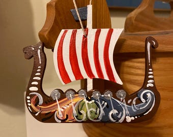 Pode incluir: Um ornamento de navio viking de madeira pintado à mão com listras vermelhas e brancas na vela e detalhes coloridos no casco.