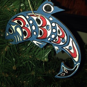 May include: A blue and red painted wooden ornament shaped like a fish with a white background and black details. The ornament is hanging from a tree branch.
