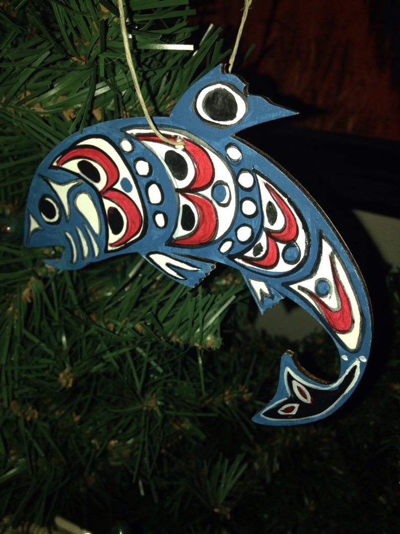 May include: A blue and red painted wooden ornament shaped like a fish with a white background and black details. The ornament is hanging from a tree branch.