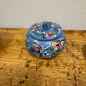 May include: A small, round, blue wooden trinket box with a lid. The box is decorated with hand-painted floral designs in pink, yellow, and white. The box sits on a wooden surface.