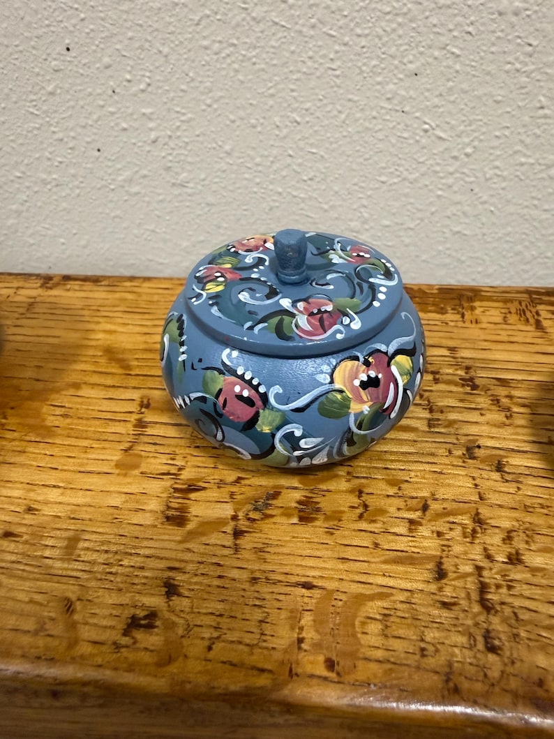 May include: A small, round, blue wooden trinket box with a lid. The box is decorated with hand-painted floral designs in pink, yellow, and white. The box sits on a wooden surface.