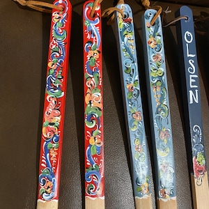 Op de afbeelding: Vijf houten lepels met handgeschilderde ontwerpen. De lepels zijn rood, blauw en wit met bloemmotieven. E&eacute;n lepel heeft het woord "OLSEN" erop geschilderd.