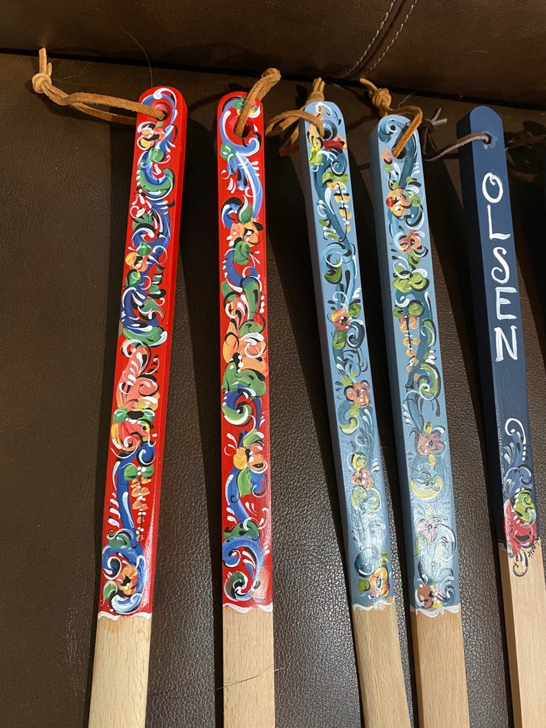 Op de afbeelding: Vijf houten lepels met handgeschilderde ontwerpen. De lepels zijn rood, blauw en wit met bloemmotieven. E&eacute;n lepel heeft het woord "OLSEN" erop geschilderd.
