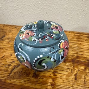 May include: A teal-colored, round wooden trinket box with a matching lid. The box and lid are adorned with hand-painted floral designs in shades of red, yellow, and white, accented by white swirls. The box sits on a wooden surface.