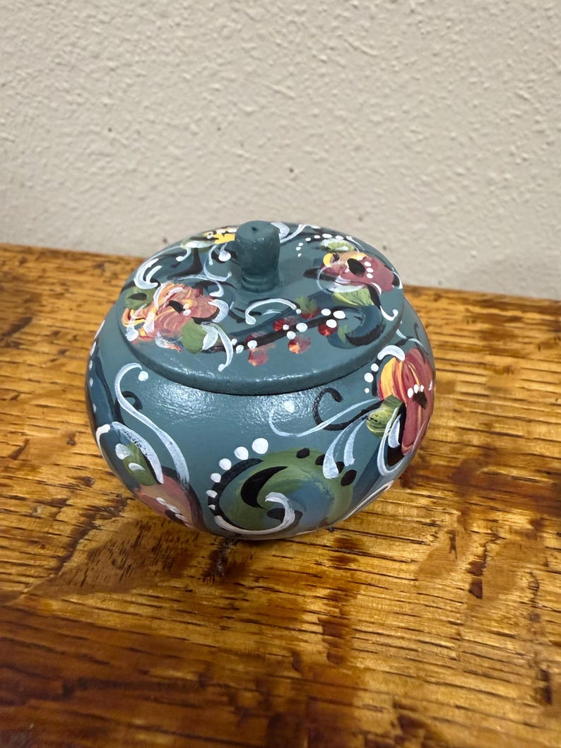 May include: A teal-colored, round wooden trinket box with a matching lid. The box and lid are adorned with hand-painted floral designs in shades of red, yellow, and white, accented by white swirls. The box sits on a wooden surface.