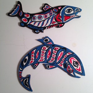 May include: Two wooden fish sculptures painted in a traditional style. The top fish is black, white, and red with a blue stripe. The bottom fish is blue, white, and red with black accents.