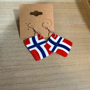 May include: A pair of earrings featuring the flag of Norway. Each earring is a red, white, and blue flag design with a silver-colored hook. The earrings are displayed on a brown earring display card.
