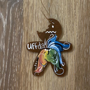 May include: A brown gingerbread person ornament with a bite taken out of it. The ornament is painted with colorful swirls and the word "Uffda!" is written on the front.