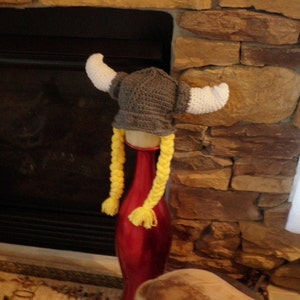 May include: A gray and white crocheted Viking hat with yellow braids. The hat is sitting on a red vase in front of a stone fireplace.