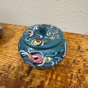 May include: A teal-colored, round wooden trinket box with a lid. The box is adorned with hand-painted floral designs in pink, white, and green. The box sits on a wooden surface.