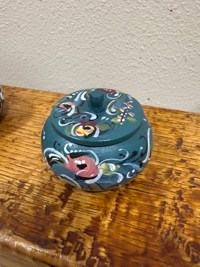 May include: A teal-colored, round wooden trinket box with a lid. The box is adorned with hand-painted floral designs in pink, white, and green. The box sits on a wooden surface.