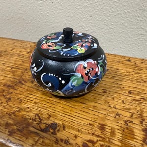 May include: A small, round, black wooden trinket box with a lid. The box and lid are decorated with colorful floral patterns in red, blue, green, and white. The box sits on a wooden surface.