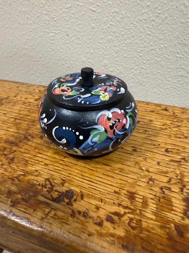May include: A small, round, black wooden trinket box with a lid. The box and lid are decorated with colorful floral patterns in red, blue, green, and white. The box sits on a wooden surface.