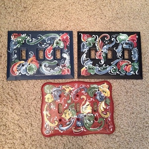 May include: Three painted light switch covers with floral designs. The top two are blue with white and red flowers. The bottom one is red with yellow, green, and white flowers.