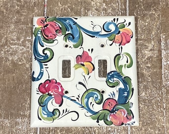 May include: A white ceramic light switch cover with a hand-painted design of blue, green, yellow, and pink flowers and swirls. The switch cover has two toggle switches.
