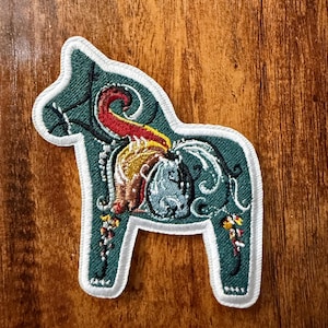 May include: An embroidered patch in the shape of a Dala horse, featuring a deep teal background with white border stitching. The design includes intricate floral and scrollwork patterns in red, yellow, and light blue thread across the body.