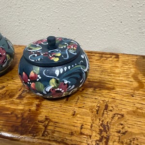 May include: A small, round, wooden trinket box with a lid. The box is painted dark blue with a floral design in red, white, yellow, and green. The box sits on a wooden surface.