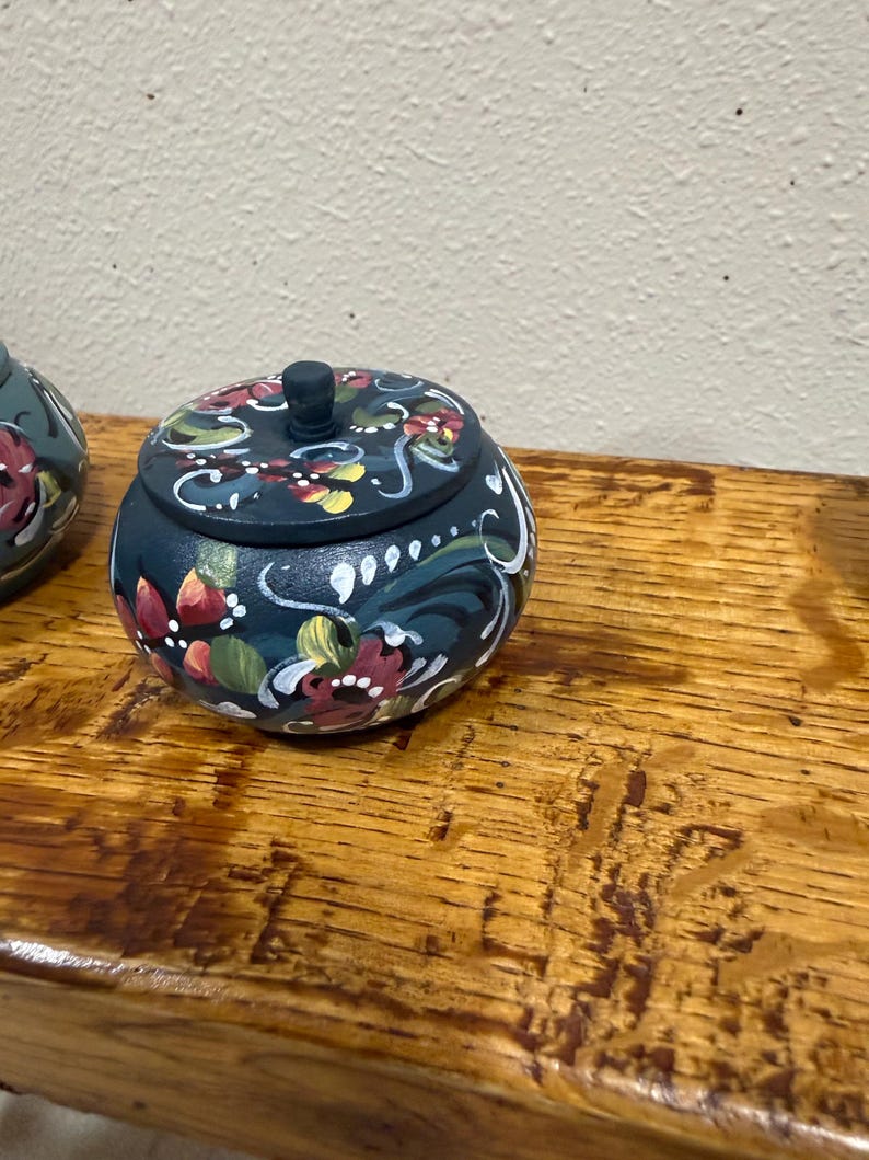 May include: A small, round, wooden trinket box with a lid. The box is painted dark blue with a floral design in red, white, yellow, and green. The box sits on a wooden surface.