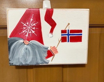 May include: A painted wooden sign featuring a gnome with a long gray beard and a red hat with a snowflake design. The gnome holds a staff with a small Danish flag. The sign has a white background.