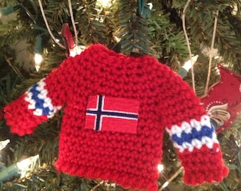 May include: A red crocheted sweater ornament with white and blue trim, featuring a Norwegian flag on the front. The ornament is hanging on a Christmas tree.