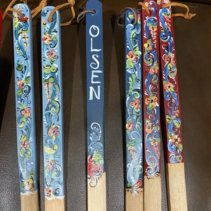Op de afbeelding: Zeven houten pinnen met beschilderde ontwerpen. De pinnen zijn beschilderd in blauw, rood en wit met bloemmotieven. E&eacute;n pin is blauw geverfd en heeft het woord "OLSEN" in wit.