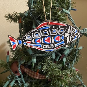 May include: A hand-painted wooden ornament shaped like a fish with black, white, red, and blue geometric patterns. The ornament is hanging on a Christmas tree.