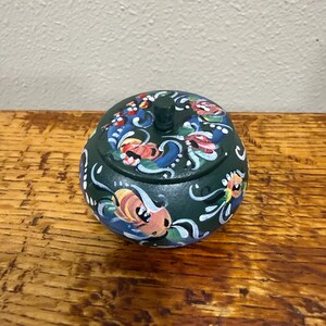 May include: A dark green, round trinket box with a lid. The box is adorned with colorful floral designs in red, blue, yellow, and white. The lid has a small, cylindrical handle. The box sits on a wooden surface.