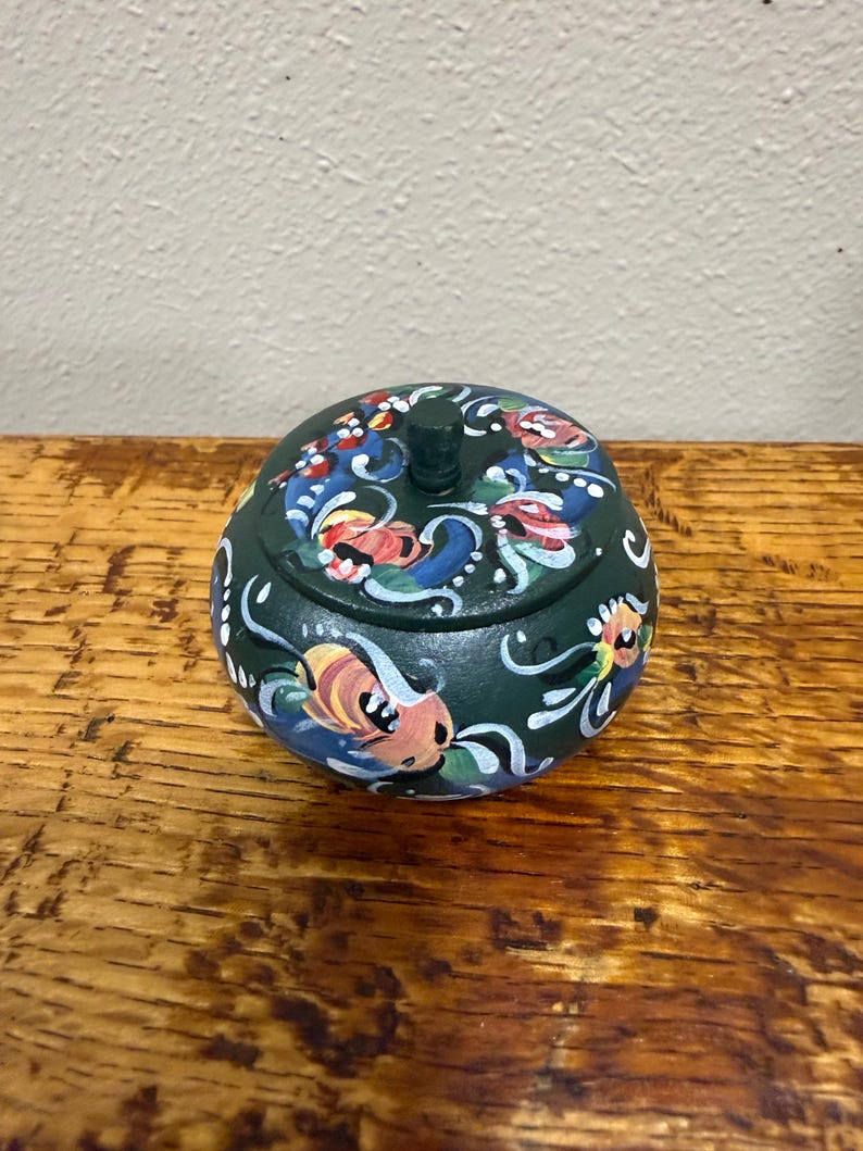 May include: A dark green, round trinket box with a lid. The box is adorned with colorful floral designs in red, blue, yellow, and white. The lid has a small, cylindrical handle. The box sits on a wooden surface.