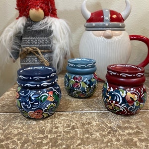 May include: Three small wooden jars with hand-painted floral designs. The jars are painted in blue, red, and gray with white accents. The jars are sitting on a wooden surface.